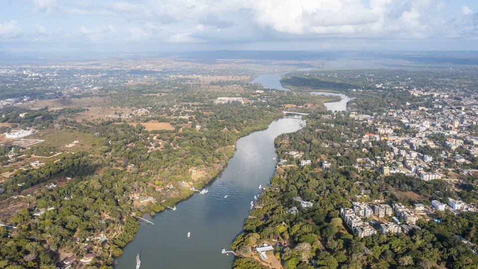 An aerial view of a river winding through a densely populated urban area with surrounding greenery and distant mountains.