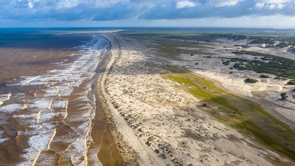 A coastal landscape with a sandy beach, ocean waves, and distant greenery.