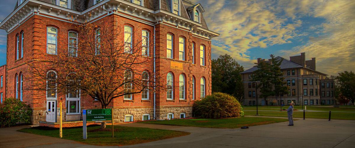 A large red brick building with arched windows and a clock tower, surrounded by a grassy area and a clear sky.