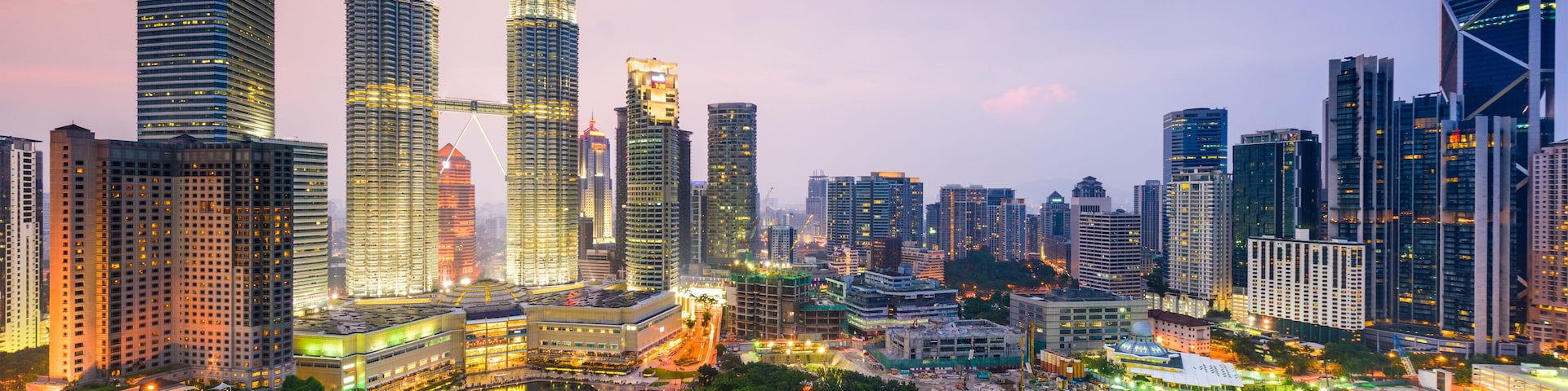 Paisagem urbana com arranha-céus imponentes, incluindo as Torres Petronas, e uma área de parque bem cuidada.