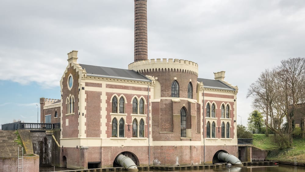 Ein historisches Backsteingebäude mit Rundbogenfenstern und einem hohen Schornstein, gelegen an einem Kanal.
