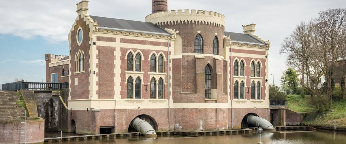 A historic brick building with arched windows and a tall chimney, situated by a canal.