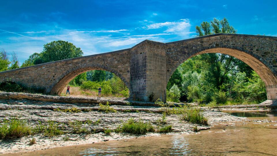 An ancient stone bridge with three arches over a river, surrounded by lush greenery and trees.