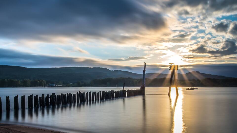 A calm lake with a wooden pier, a lighthouse, and a sunset.