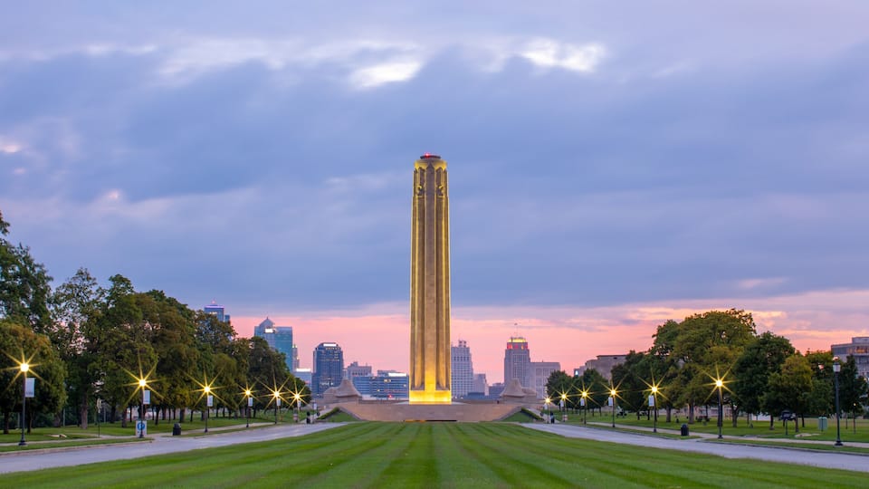 A tall illuminated monument in a park with a city skyline in the background.
