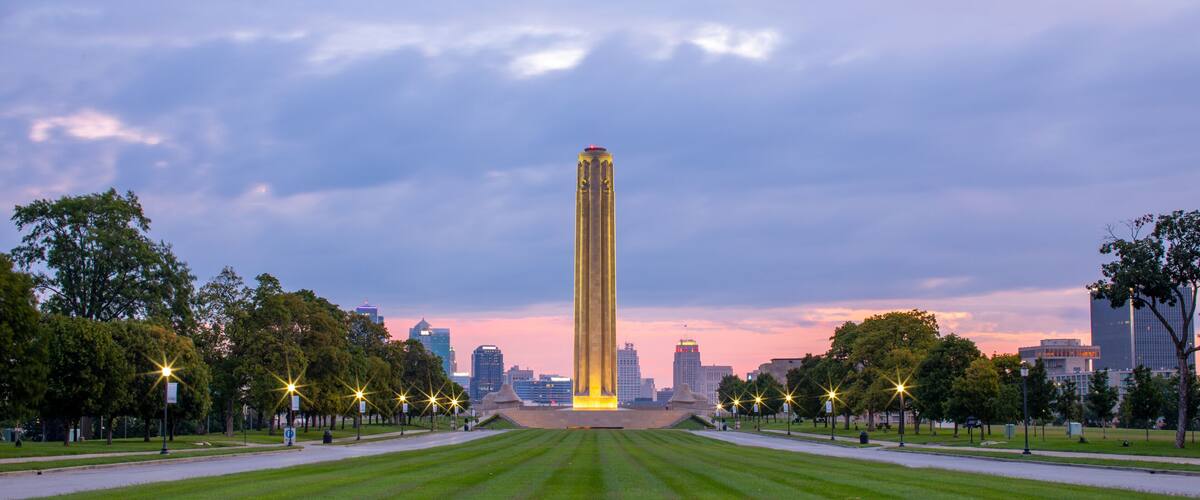 A tall illuminated monument in a park with a city skyline in the background.