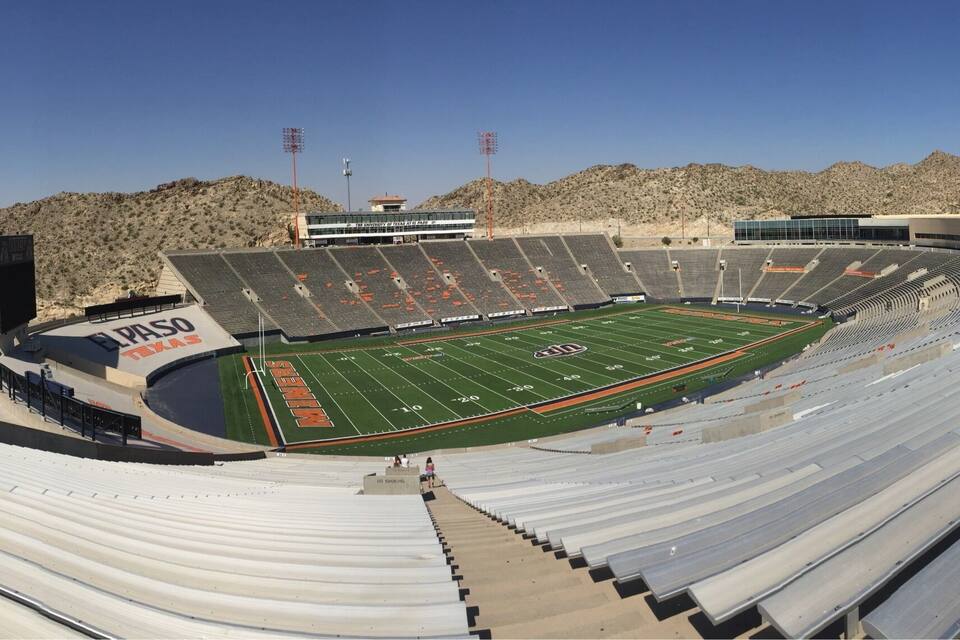 A large, empty stadium with a football field, surrounded by hills and mountains.