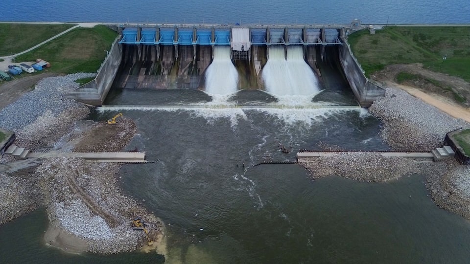 A dam with water flowing through it, a clear sky, and a calm body of water in the background.