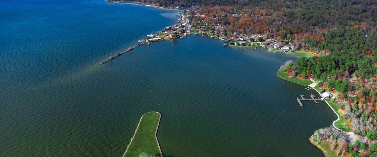 An aerial view of a lake with a dock, surrounded by dense forest and residential areas.