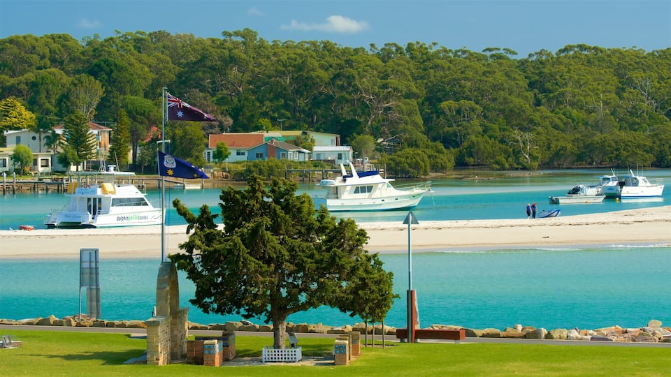 A coastal town with a clear blue sea, boats, a sandy beach, and lush green hills in the background.