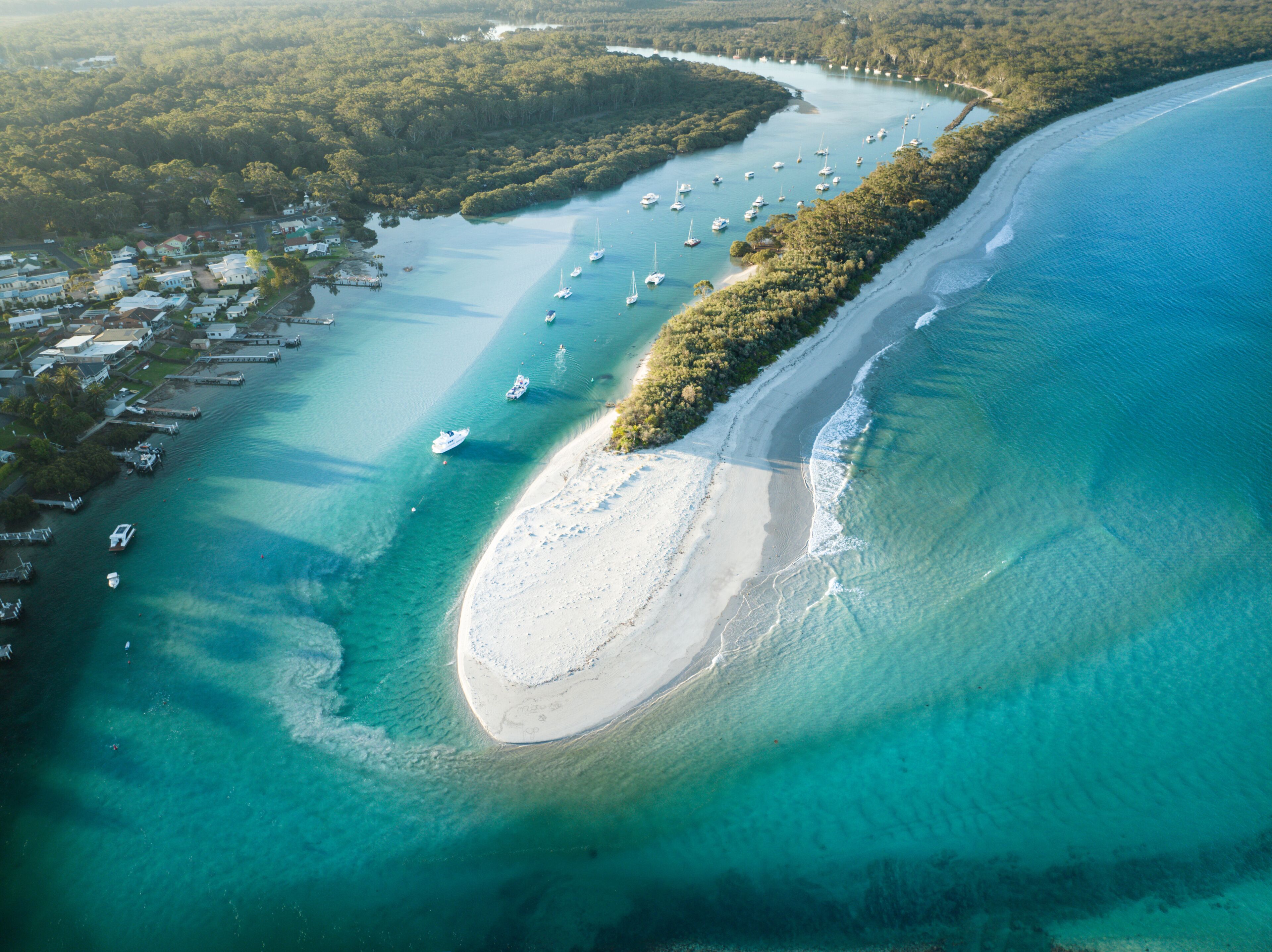 An aerial view of a coastal area with a sandy beach, clear turquoise waters, and numerous boats anchored near the shore.