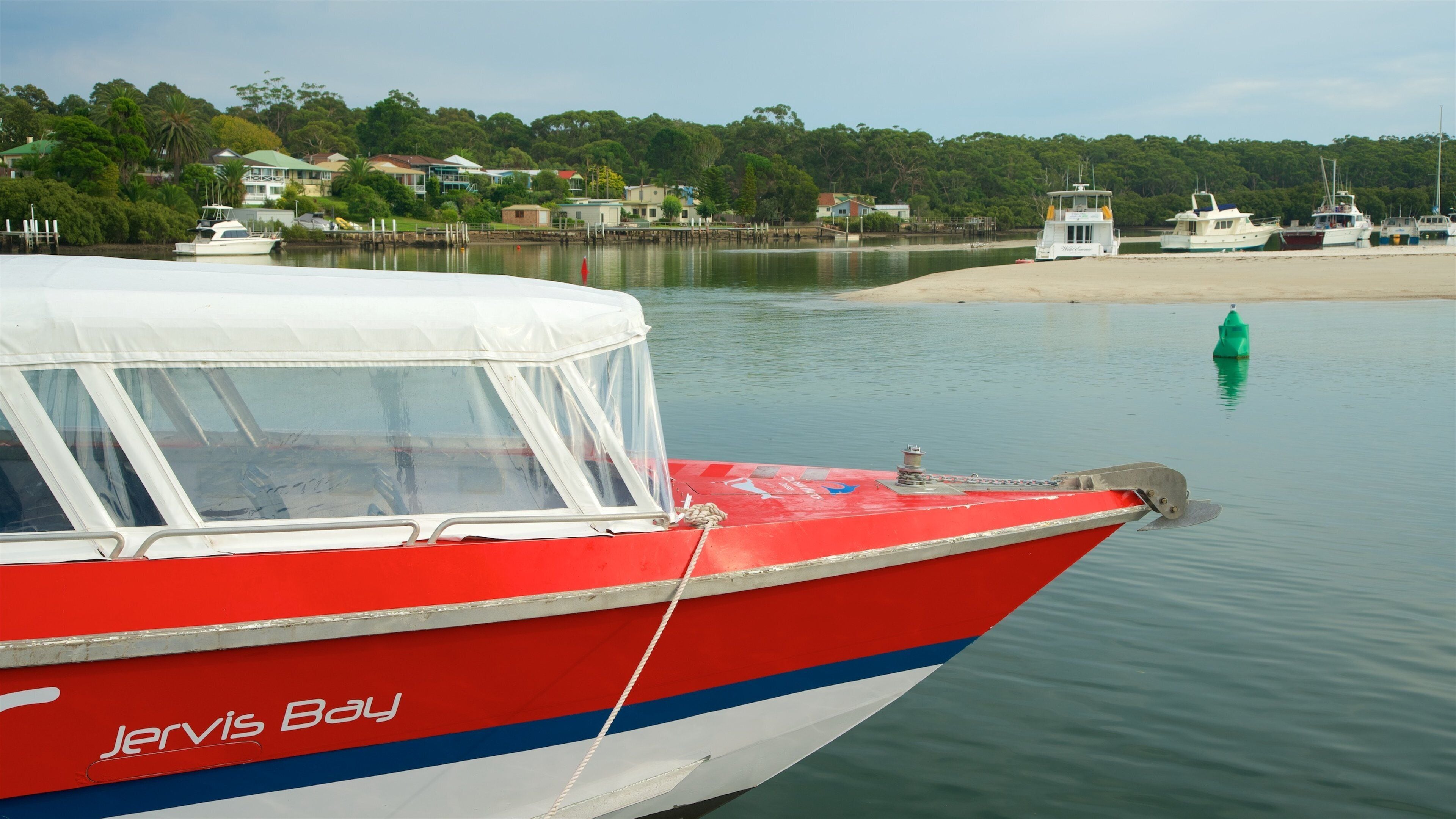 A red and white boat named 'Jervis Bay' docked at a calm waterfront with houses and greenery in the background.