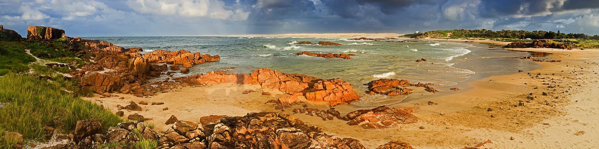 A coastal landscape with rocky outcrops, a sandy beach, and a cloudy sky.