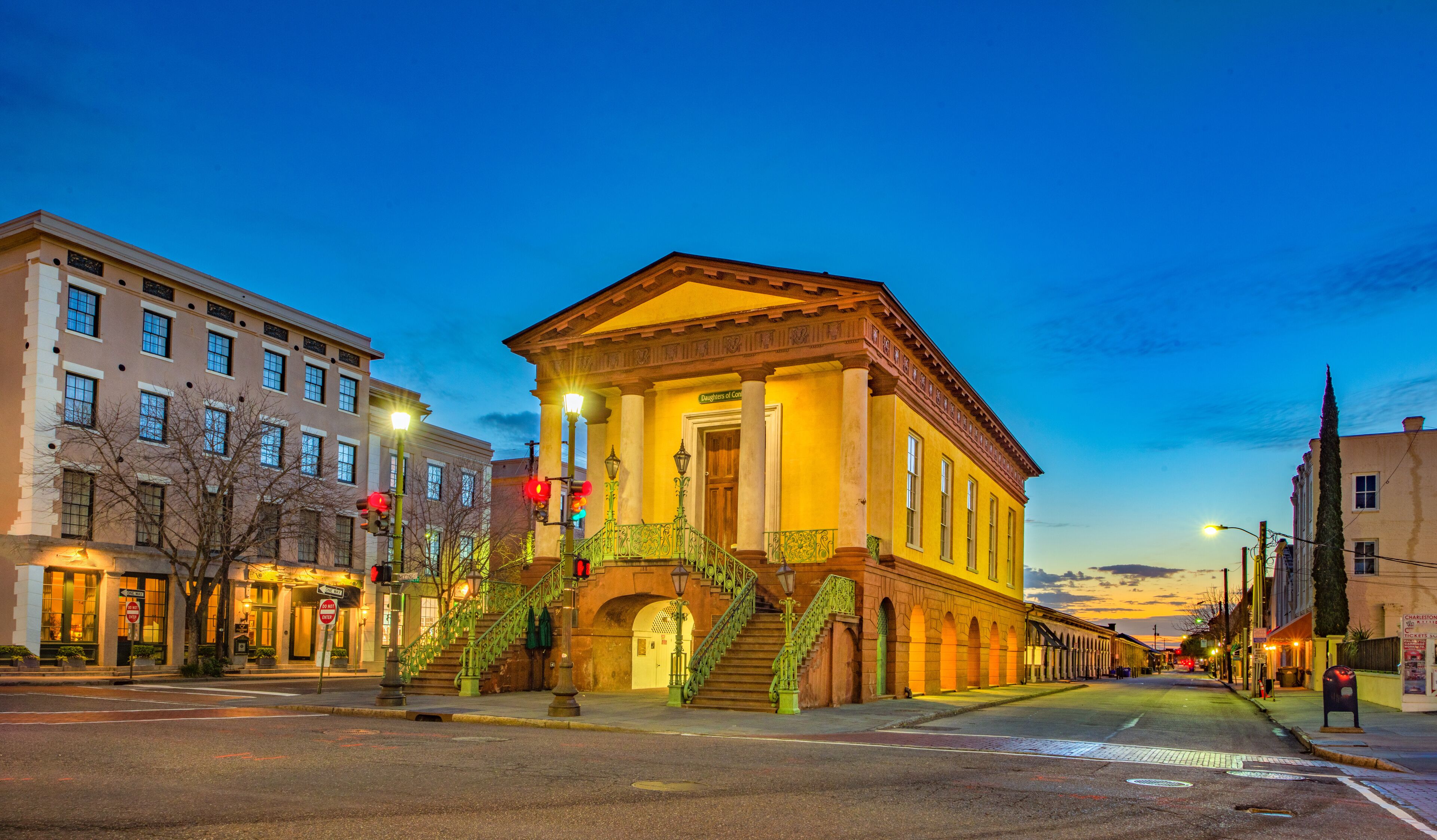 A historic building with a yellow facade and green stairs, surrounded by modern buildings and a clear sky.