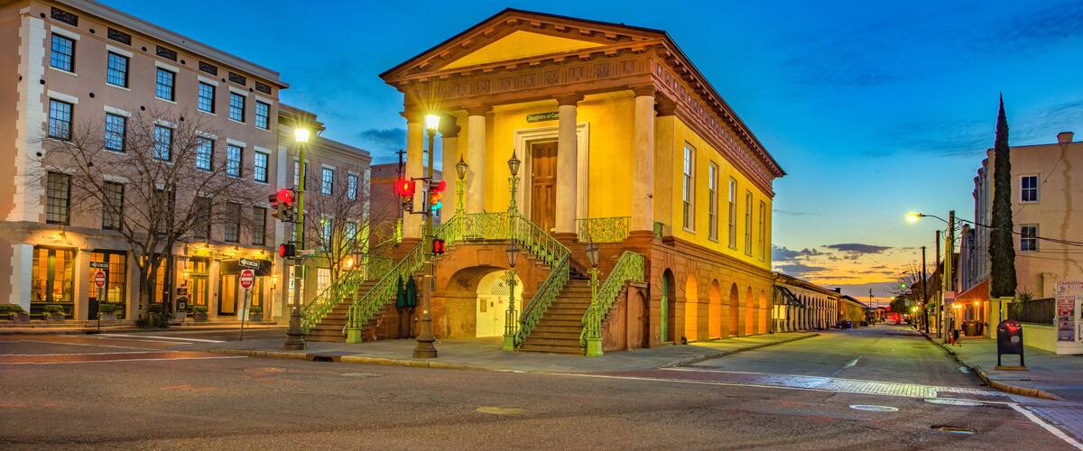 A historic building with a yellow facade and green stairs, surrounded by modern buildings and a clear sky.