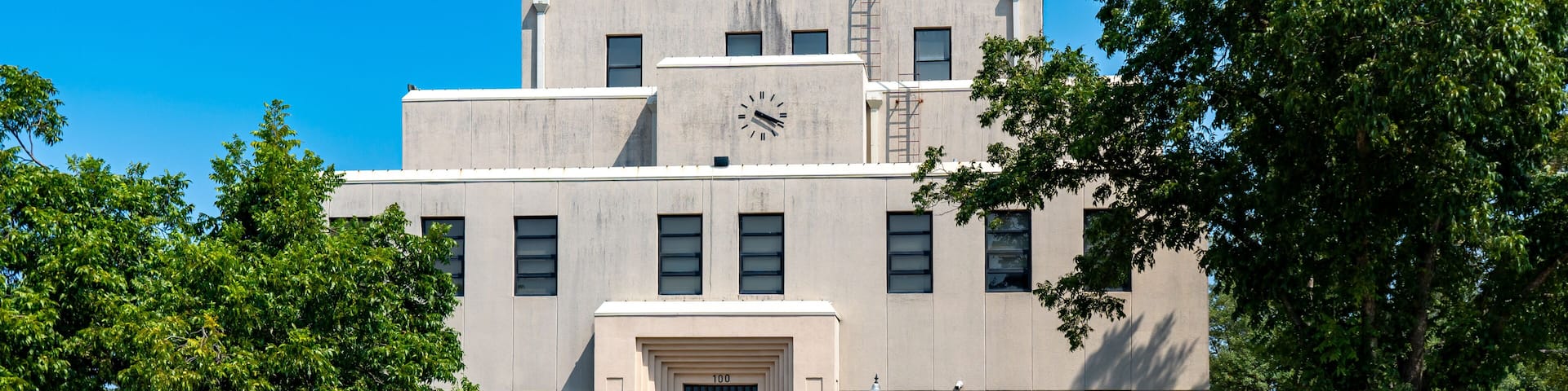 A multi-story building with a clock on the facade, a set of stairs leading to the entrance, and surrounding greenery.