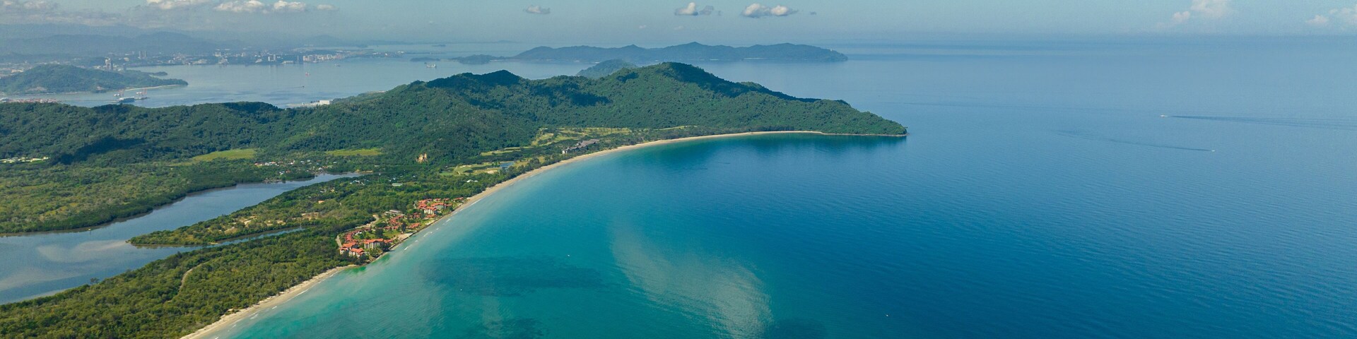 Vista aérea de una zona costera con mar azul cristalino, una playa arenosa y colinas verdes.