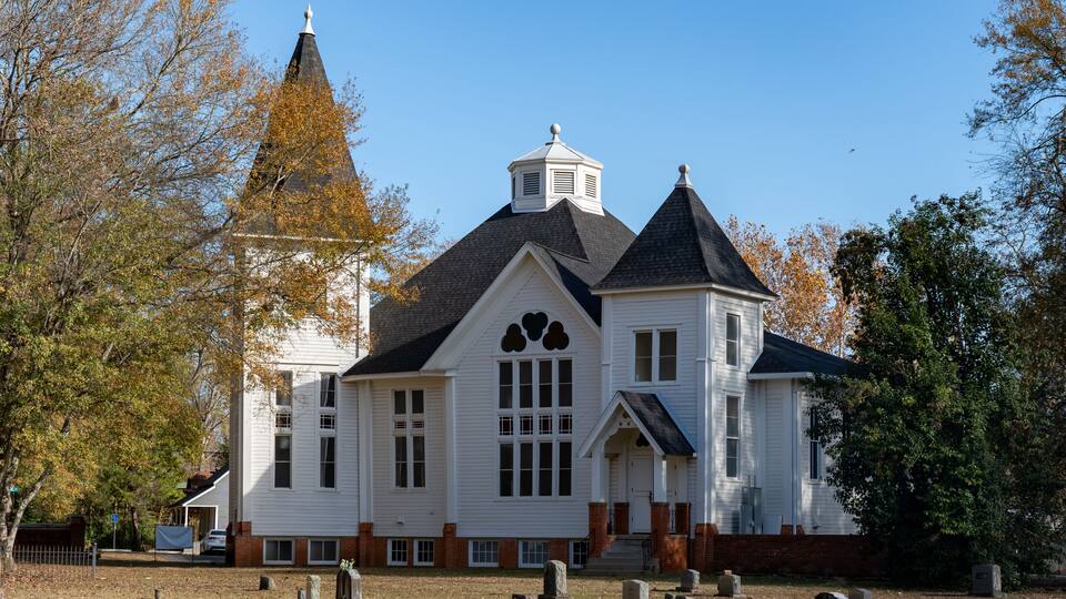A white church with a steeple and a bell tower, surrounded by trees and gravestones.