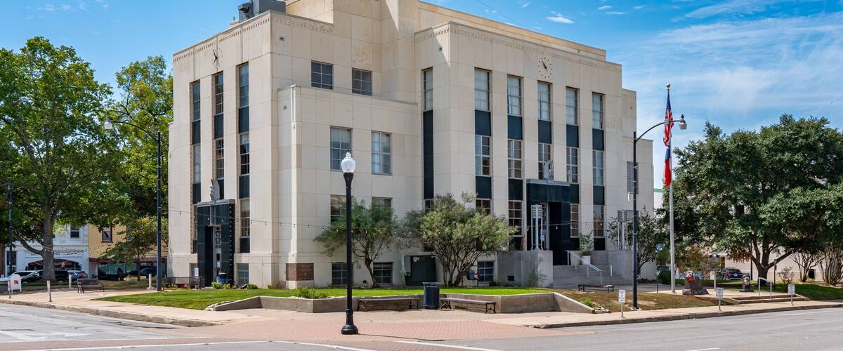 A large beige building with multiple windows, a flagpole with an American flag, and a clear blue sky with scattered clouds.