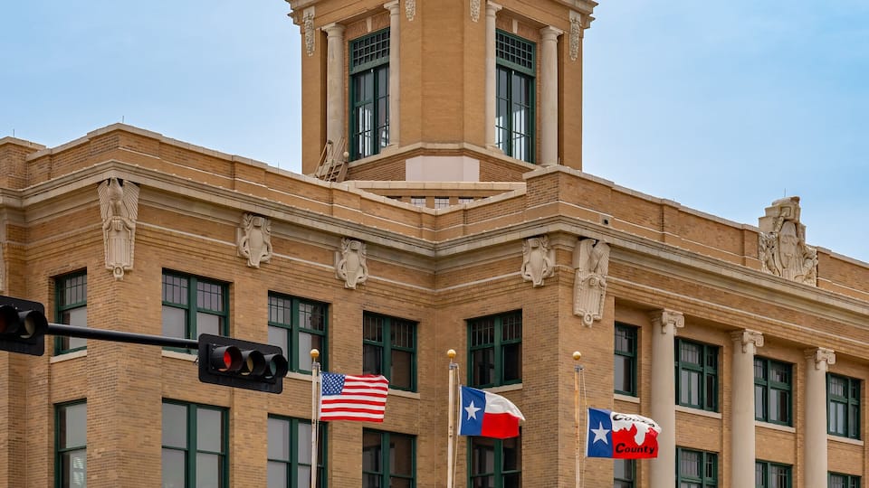 A historic building with a dome and clock tower, featuring multiple flags including the Texas state flag.