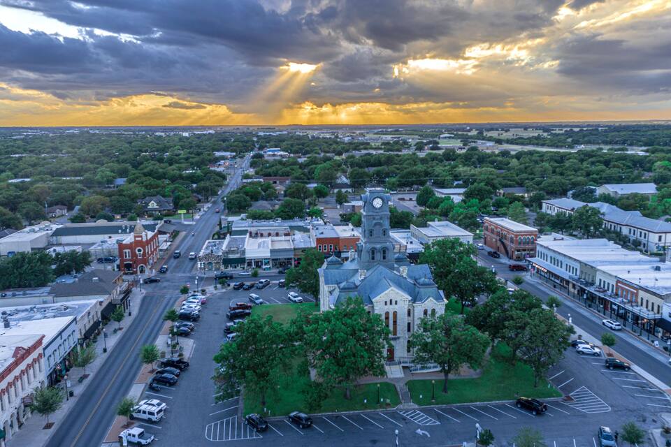 A town square with a prominent clock tower, surrounded by historic buildings and a large parking lot.