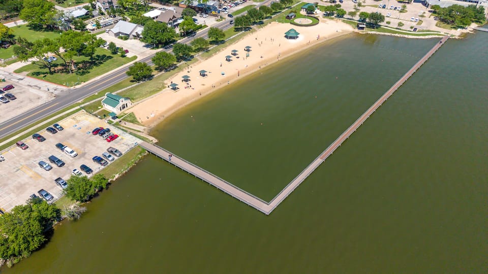 An aerial view of a lake with a dock, a parking lot, and surrounding buildings.