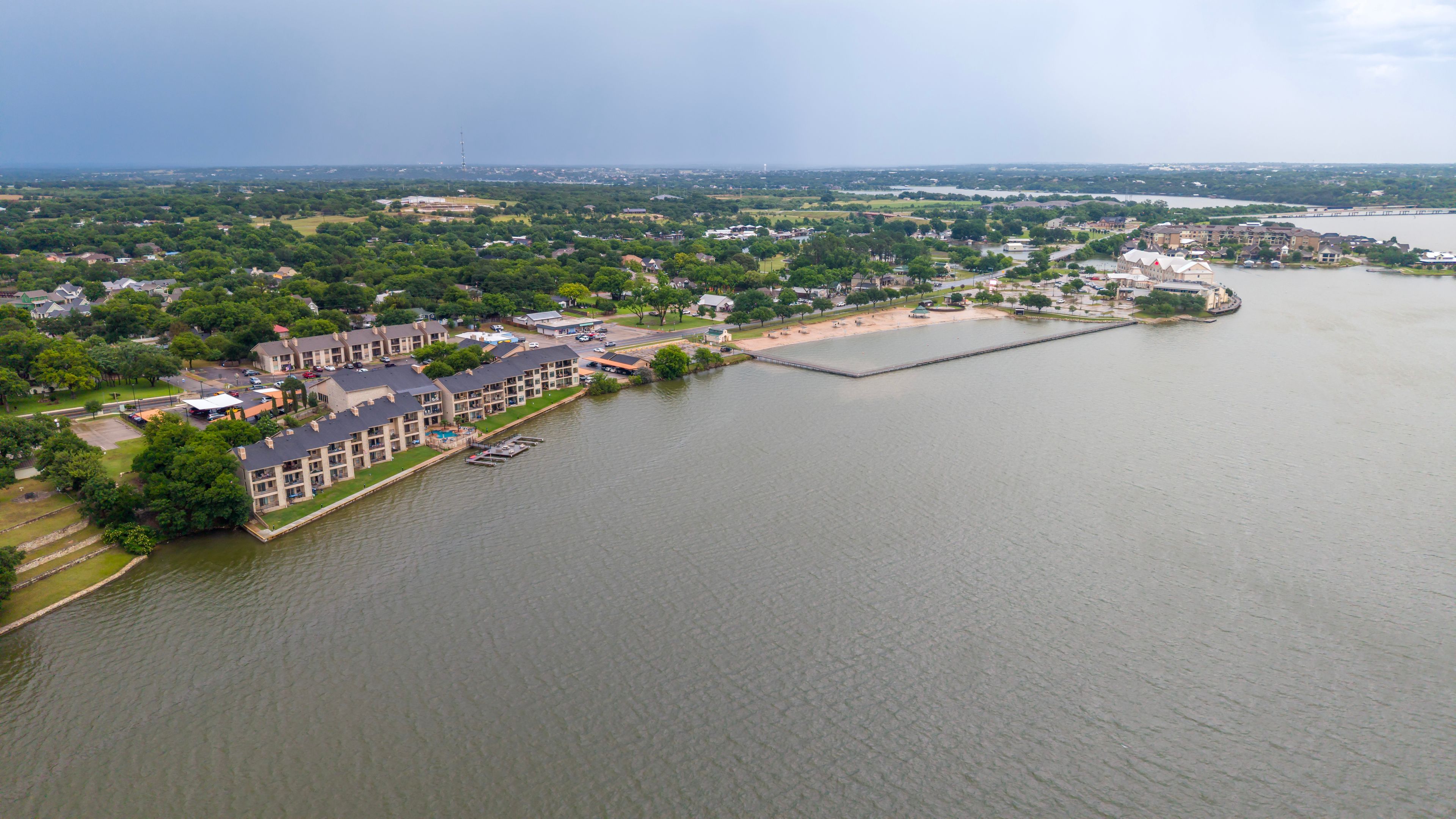 An aerial view of a riverside residential area with a dock, a bridge in the distance, and a large body of water.
