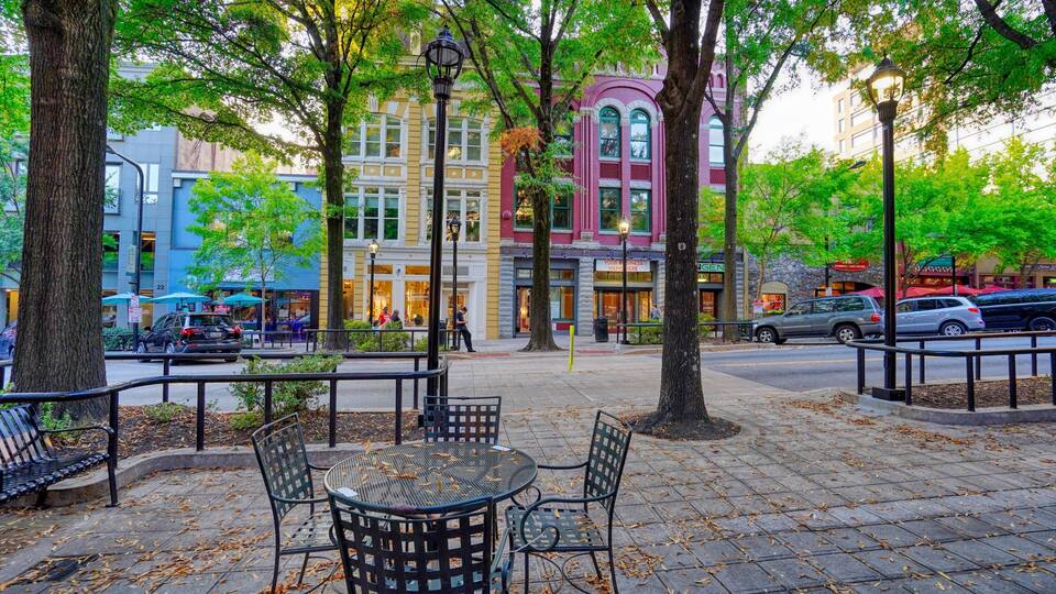 A park with a metal table and chairs, trees, and buildings with storefronts.