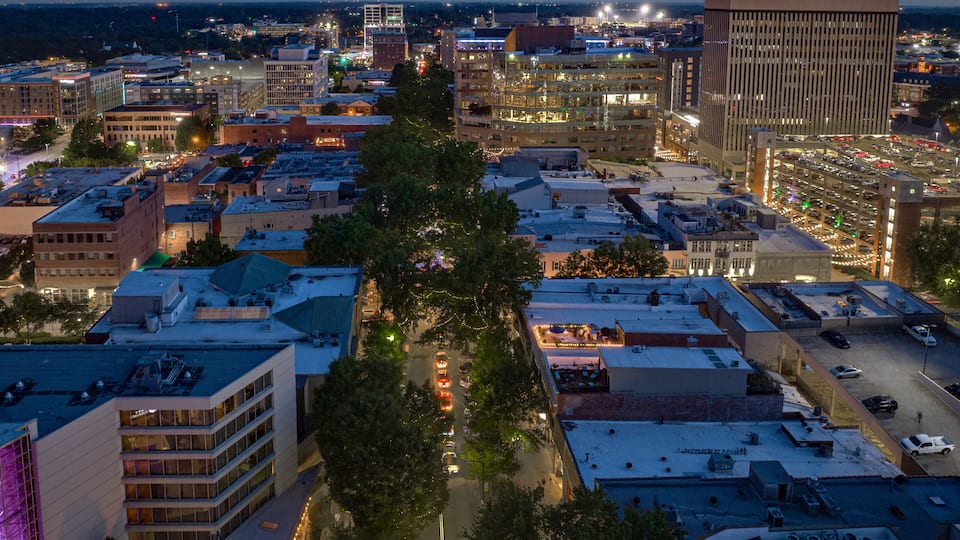 A cityscape at dusk with illuminated buildings, a central tree-lined street, and a parking structure.