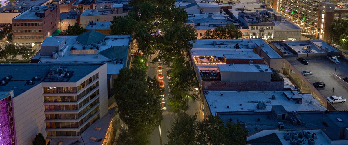 A cityscape at dusk with illuminated buildings, a central tree-lined street, and a parking structure.