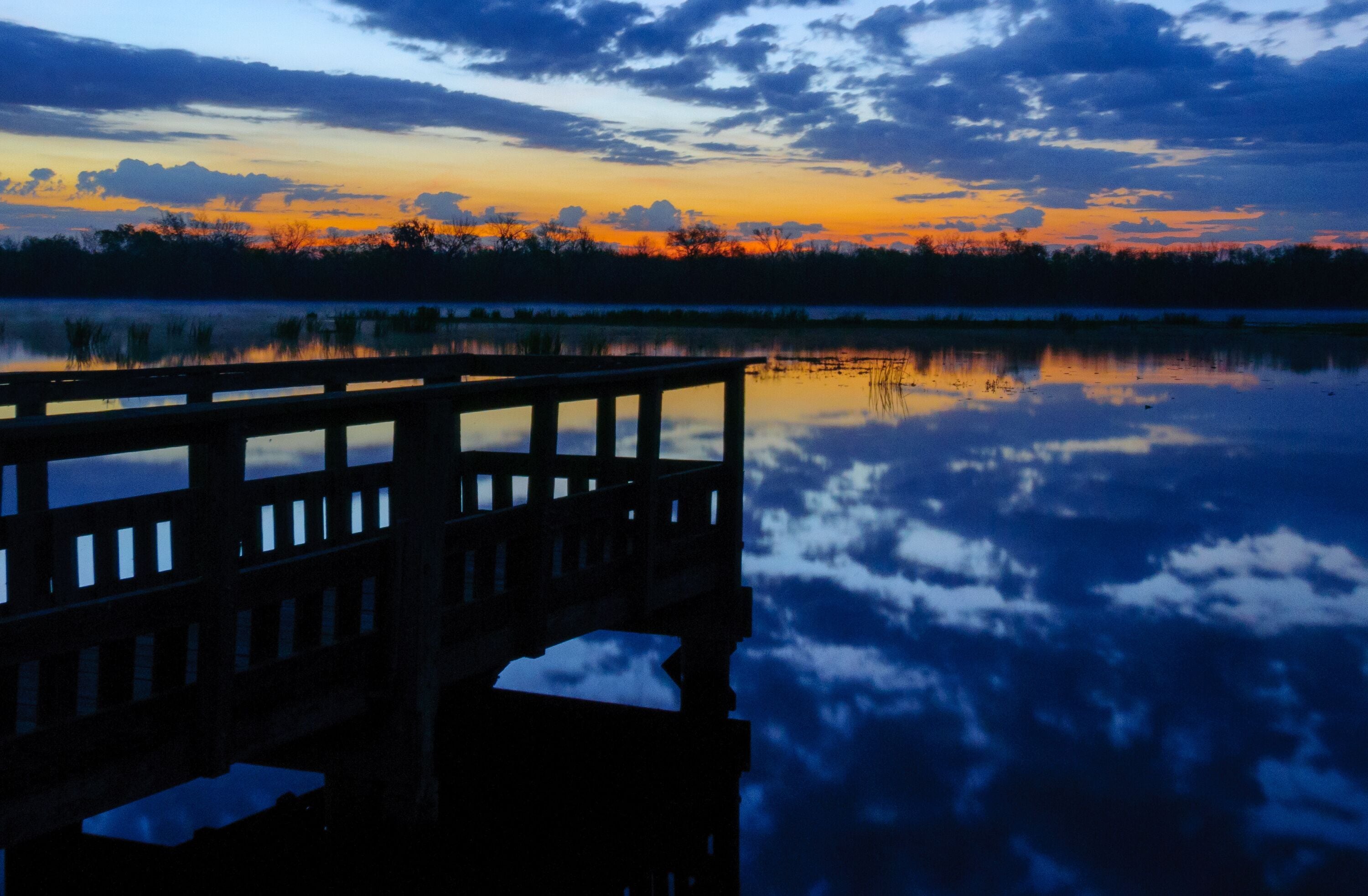 Pôr do sol sobre um lago calmo com uma ponte de madeira refletindo o céu.