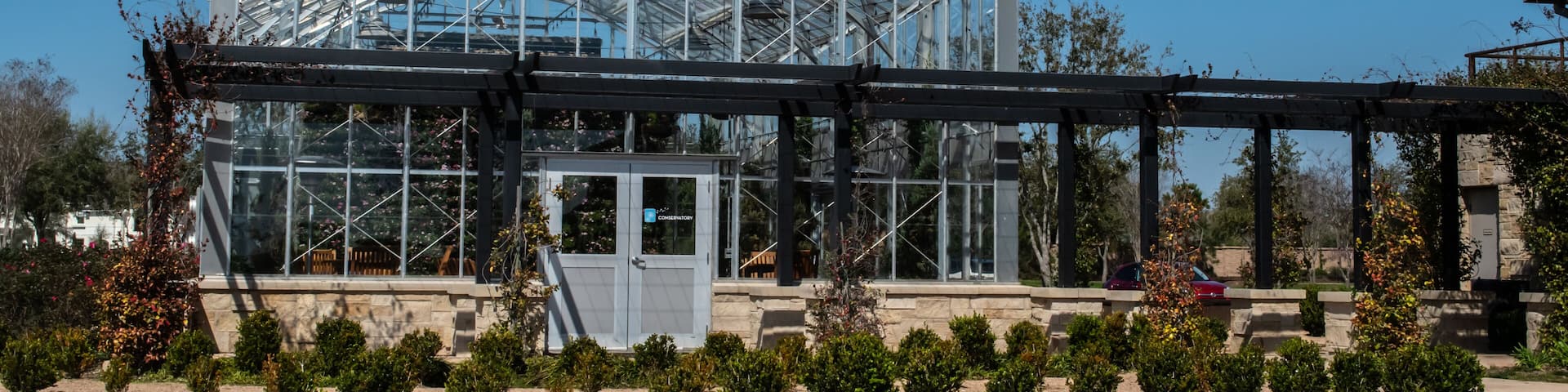 A large glass greenhouse structure with a metal frame, surrounded by a landscaped garden with small bushes and a clear blue sky.