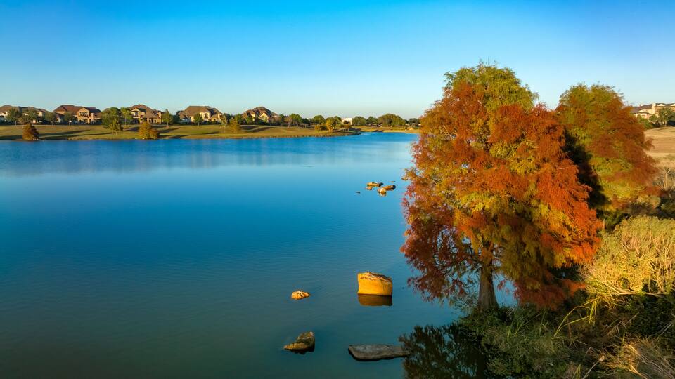 A calm lake with autumn-colored trees and houses in the background.
