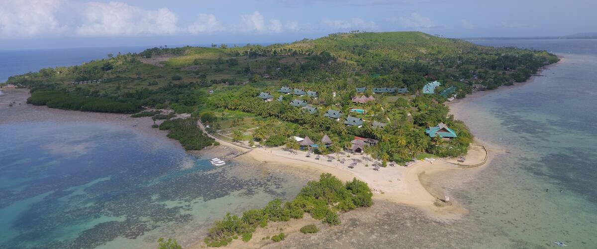 An island with a beach, clear waters, and several buildings with green roofs.