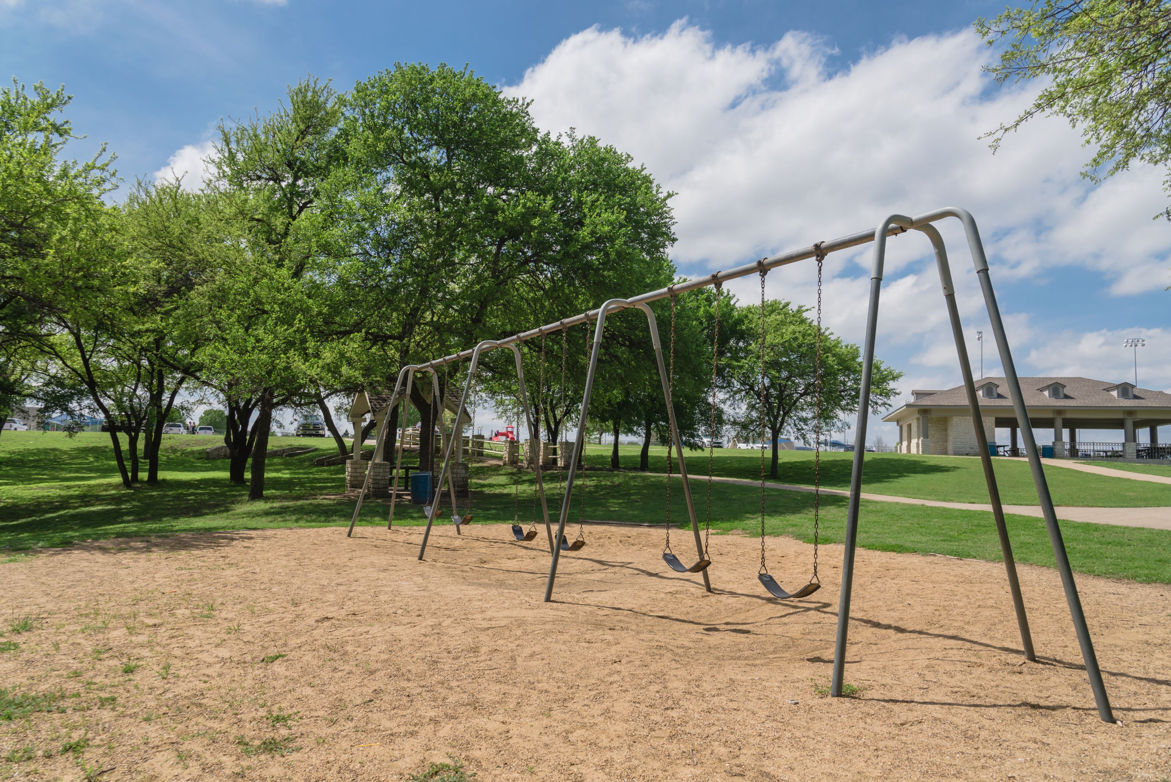 Close-up swing set 6 seats in nature park and tree lush at Ennis, Texas, USA. Outdoor sand playground hanging seat suspended from bar swings back and forth surround by leaves green, cloud blue sky
