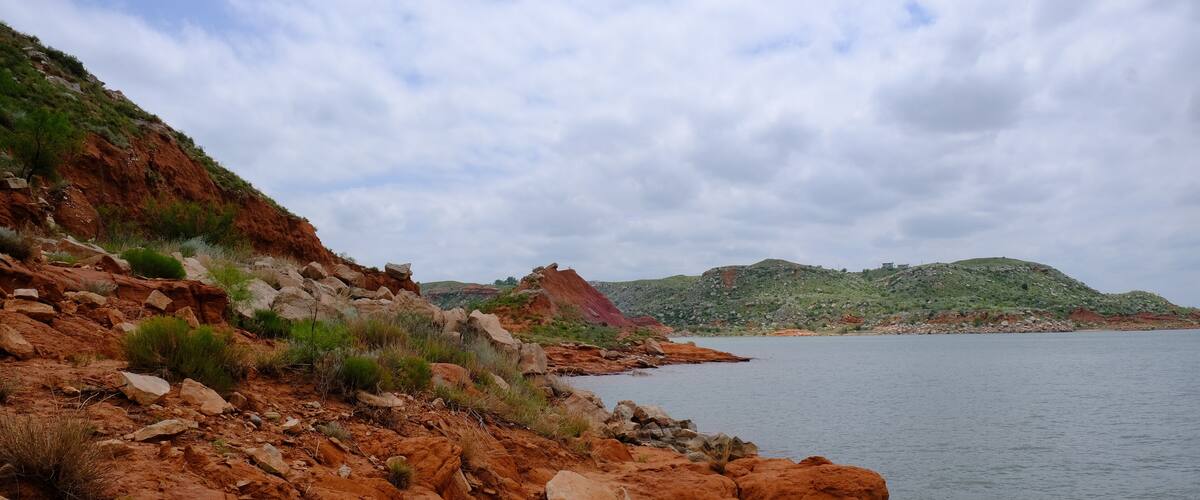 A rocky shoreline with a calm body of water, green vegetation, and distant hills.