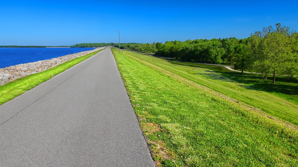 A paved path alongside a body of water, bordered by a rocky shoreline and a grassy area with trees.