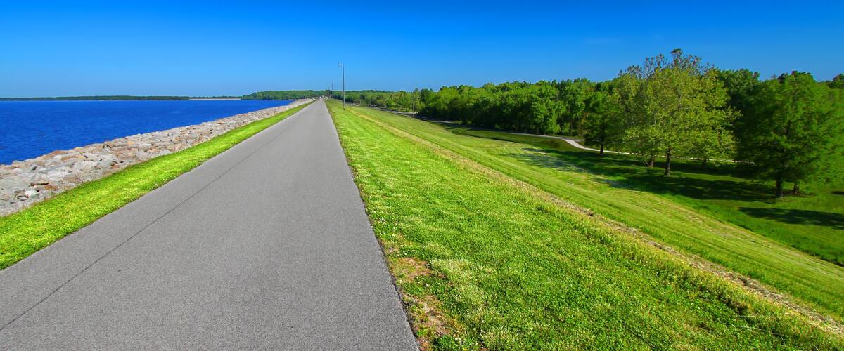 A paved path alongside a body of water, bordered by a rocky shoreline and a grassy area with trees.