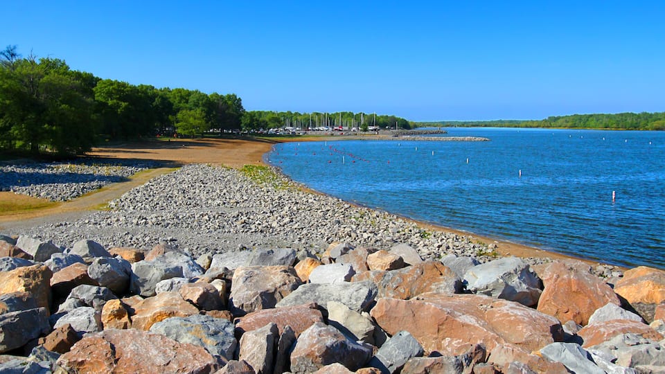 A rocky shoreline with a clear blue lake, surrounded by trees and a distant shoreline with buildings.