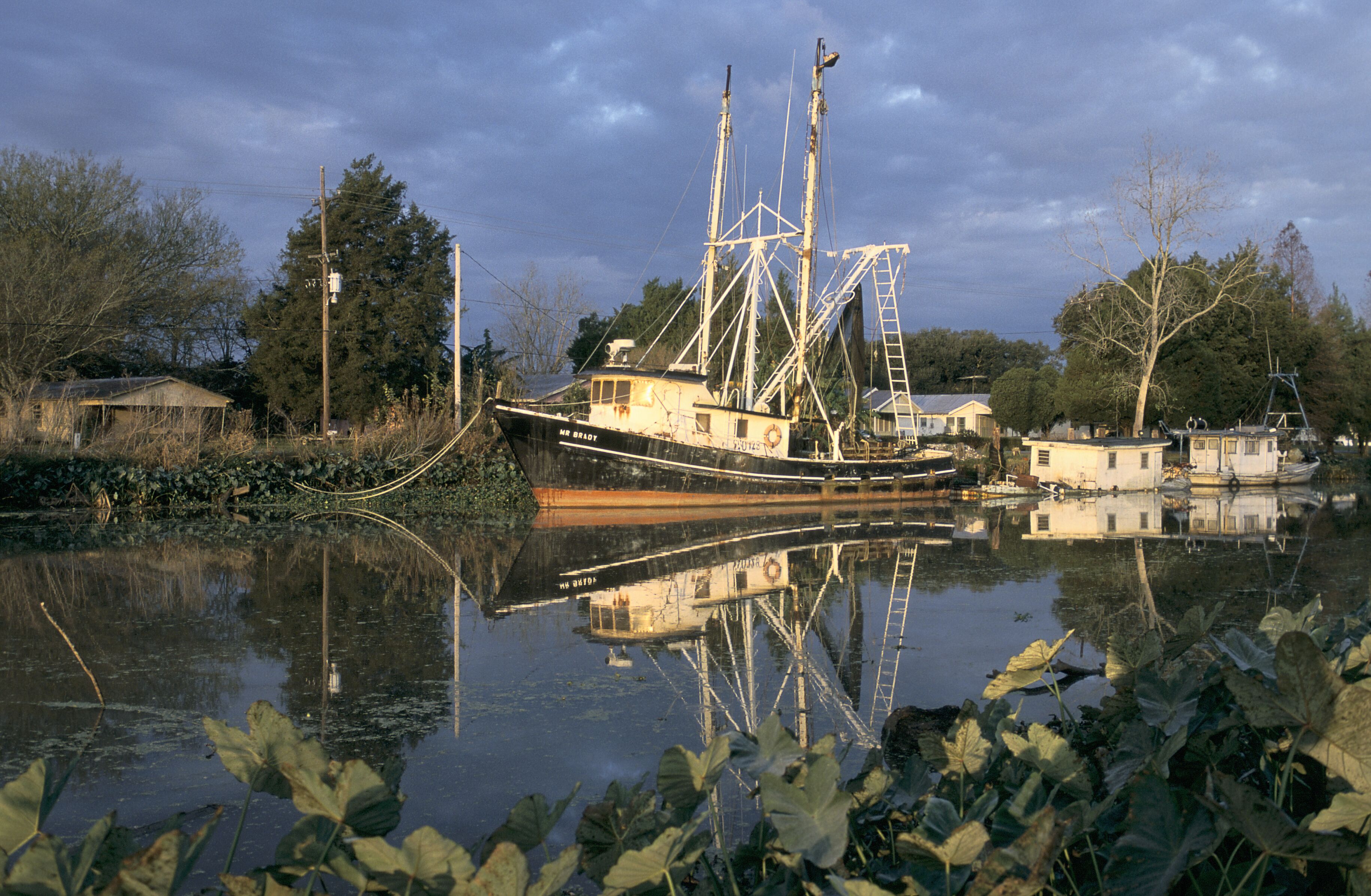A large fishing boat docked at a pier with a house and smaller boats nearby.