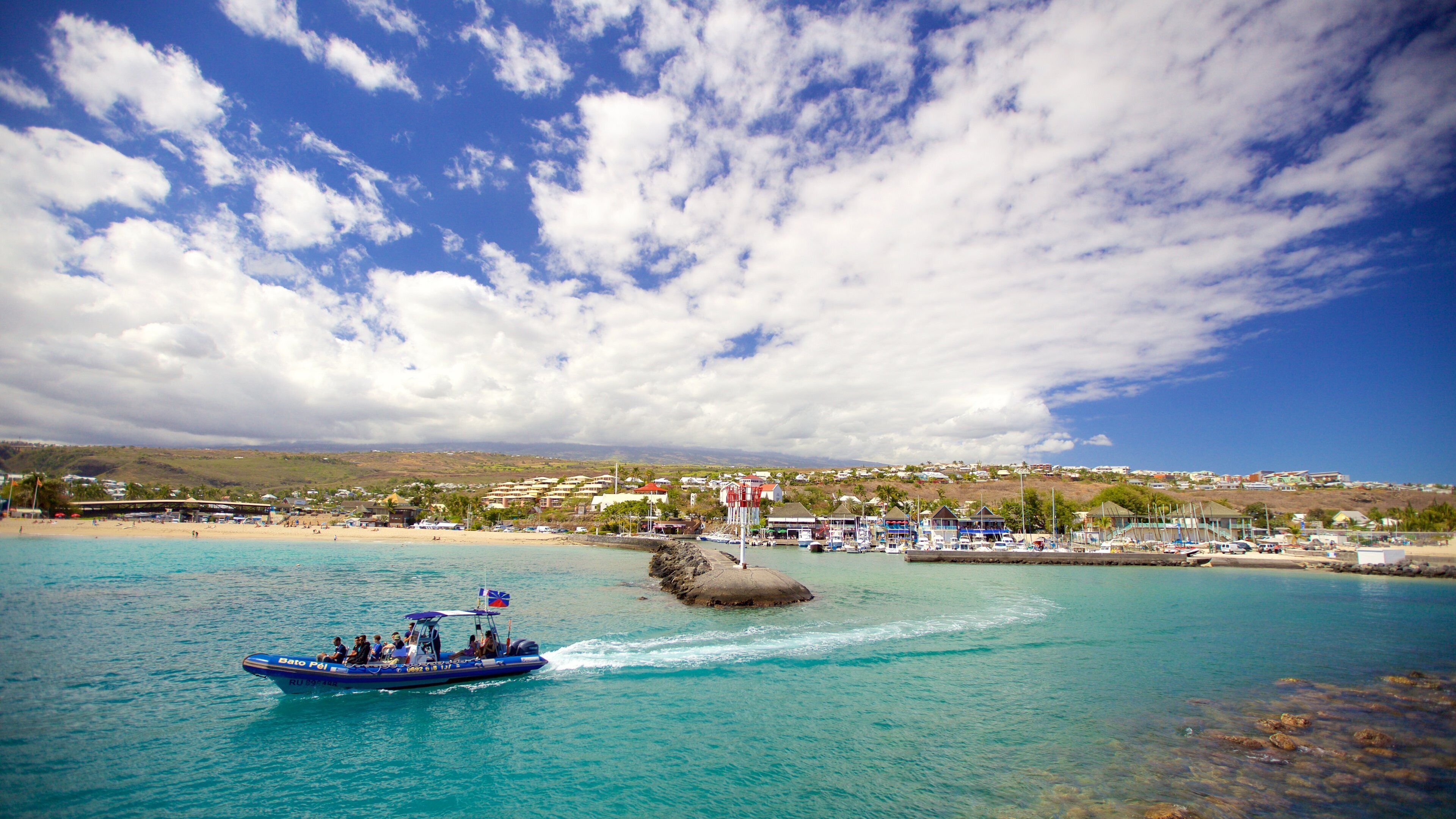 A boat on turquoise waters near a coastal town with buildings and a rocky outcrop.