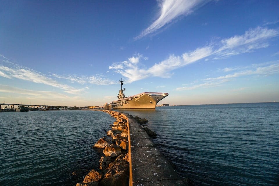 A large naval ship docked at a pier with a clear sky and calm sea.