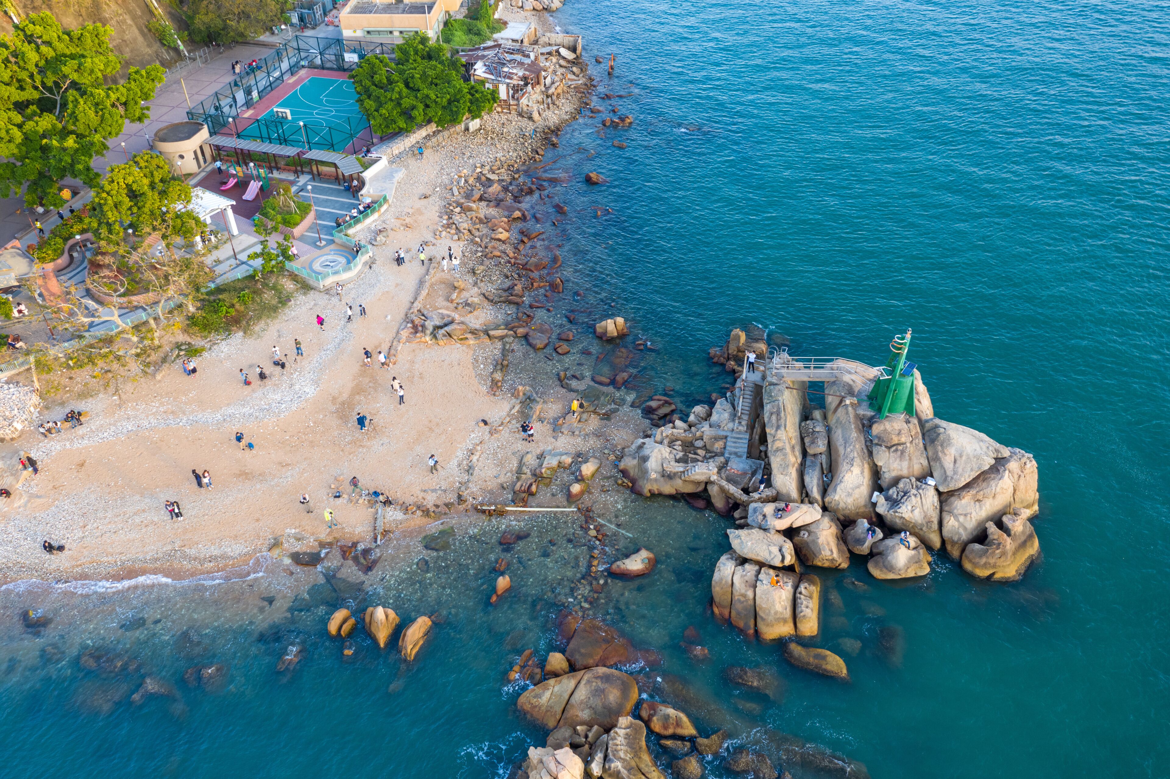 Ein Luftbild zeigt einen Strand mit Personen, eine felsige Küstenlinie und ein Gebäude mit einem Pool.