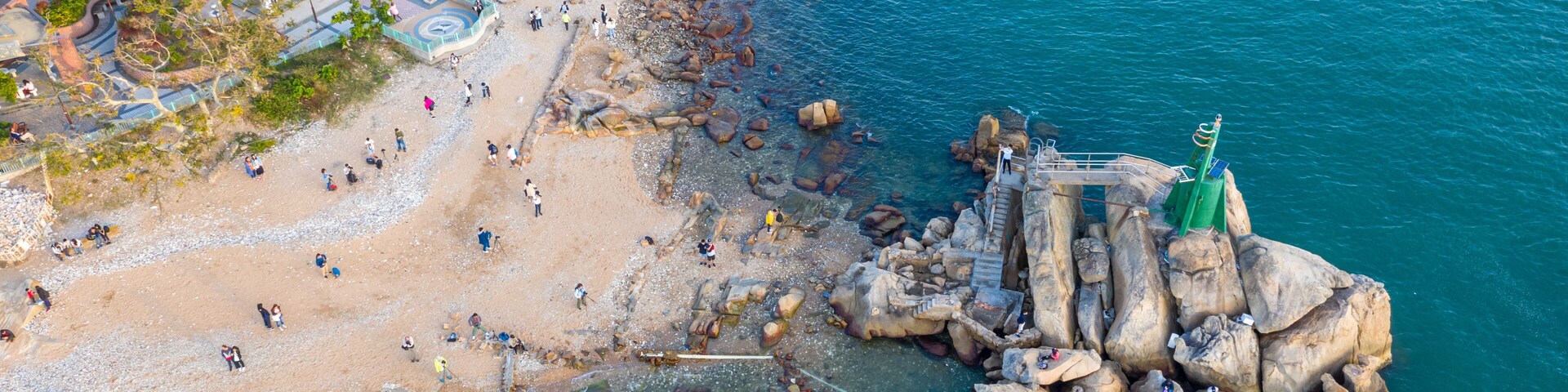 Ein Luftbild zeigt einen Strand mit Personen, eine felsige Küstenlinie und ein Gebäude mit einem Pool.