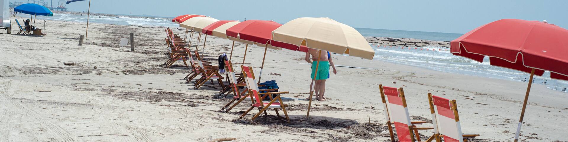 Panorama view tourist couple after row of colorful rental umbrella capture managed beach areas community-friendly coastal recreation along quiet sandy shoreline, rocky pier in Galveston, Texas
