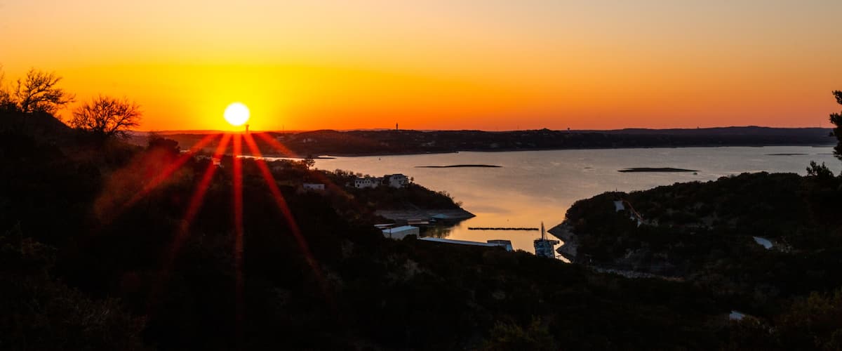 A sunset over a body of water with a silhouette of a town and hills in the background.
