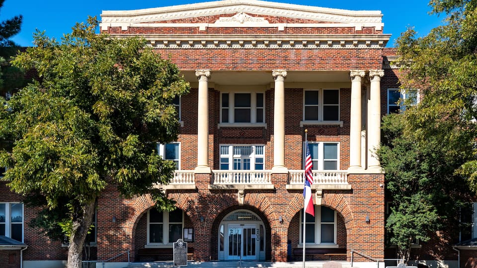 A historic brick building with columns, a flag, and a sign indicating it's a county courthouse.
