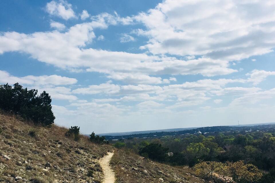 A dirt path winding through a dry, rocky landscape with sparse vegetation and a clear blue sky with scattered clouds.
