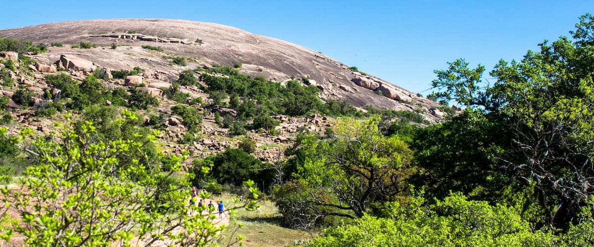A large rock formation with a clear sky, surrounded by green vegetation and a few people hiking.