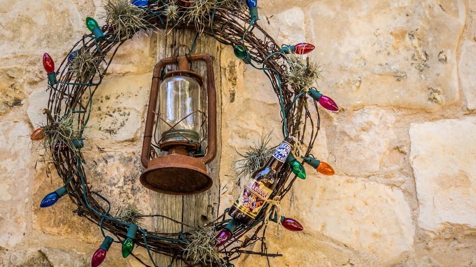 A wreath with colorful lights and a bottle of beer against a stone wall.